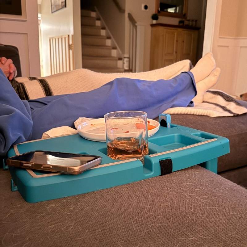 Person lounging on a couch with a stupid car tray holding a glass, phone, and plate.